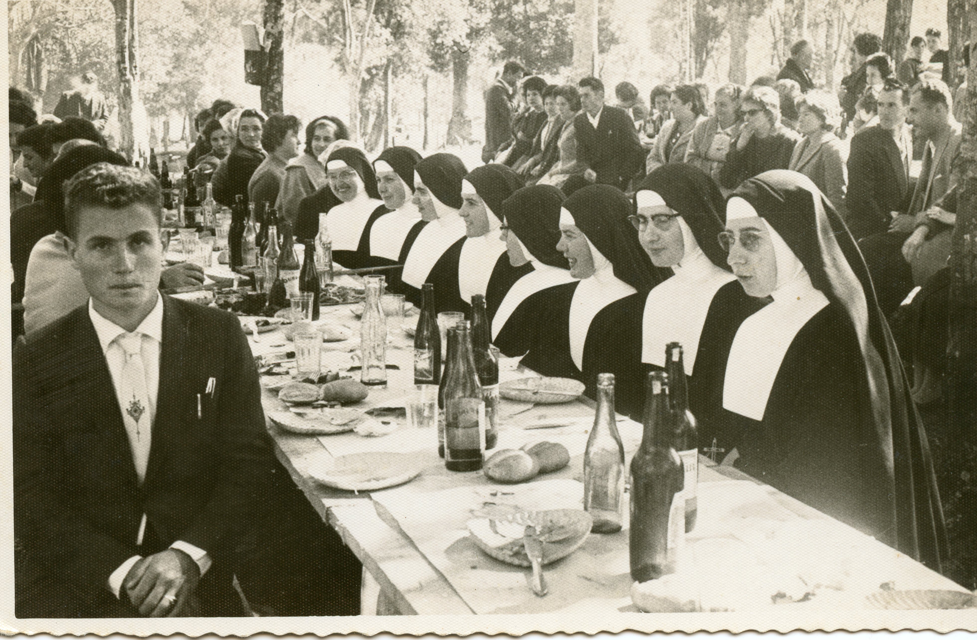Uma fotografia em preto e branco que retrata um banquete ao ar livre, possivelmente um evento comunitário ou religioso, com um grupo de freiras sentadas à mesa e um homem em primeiro plano. A cena sugere um contexto social de meados do século XX, com vestimentas formais e hábitos religiosos tradicionais, em um ambiente arborizado que remete a festividades típicas de pequenas cidades brasileiras.