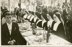 Uma fotografia em preto e branco que retrata um banquete ao ar livre, possivelmente um evento comunitário ou religioso, com um grupo de freiras sentadas à mesa e um homem em primeiro plano. A cena sugere um contexto social de meados do século XX, com vestimentas formais e hábitos religiosos tradicionais, em um ambiente arborizado que remete a festividades típicas de pequenas cidades brasileiras.