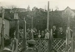 Fotografia em preto e branco de uma cerimônia cívica ao ar livre, possivelmente em uma pequena cidade brasileira, com autoridades e uma mulher discursando em um palanque de madeira. O cenário inclui bandeiras do Brasil e construções típicas de colonização europeia ao fundo, sugerindo um evento oficial ou comemorativo de meados do século XX.