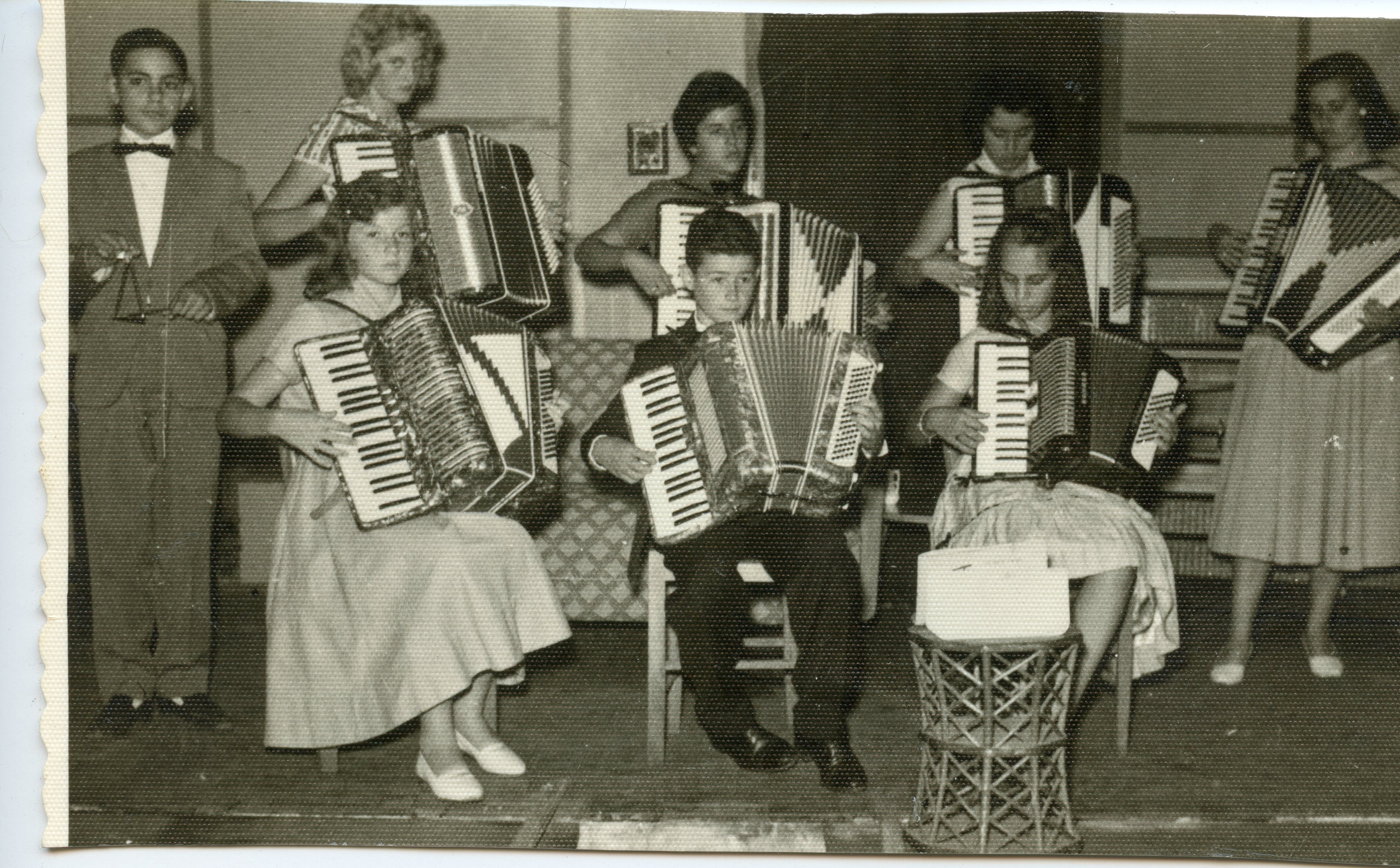 Uma fotografia em preto e branco retratando um grupo de jovens músicos tocando acordeons em um ambiente interno, possivelmente um palco ou salão de eventos. O grupo, composto por meninos e meninas vestidos formalmente, sugere uma apresentação musical escolar ou comunitária típica de meados do século XX.