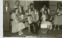 Uma fotografia em preto e branco retratando um grupo de jovens músicos tocando acordeons em um ambiente interno, possivelmente um palco ou salão de eventos. O grupo, composto por meninos e meninas vestidos formalmente, sugere uma apresentação musical escolar ou comunitária típica de meados do século XX.