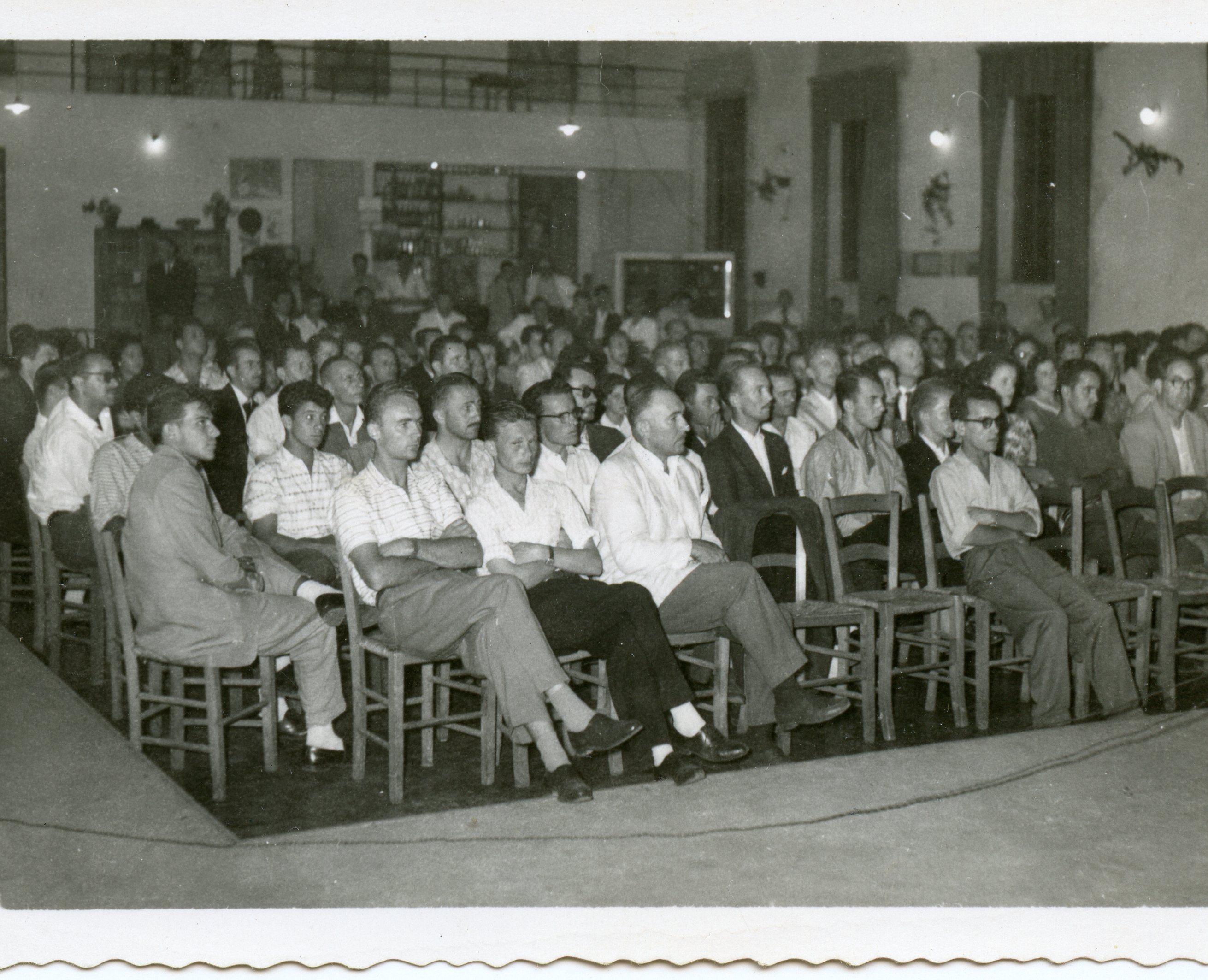 Uma fotografia em preto e branco que retrata uma plateia composta majoritariamente por homens sentados em cadeiras de madeira em um salão fechado. O ambiente sugere uma reunião, palestra ou evento comunitário, com vestimentas típicas das décadas de 1950 ou 1960, como camisas sociais e paletós.