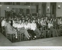 Uma fotografia em preto e branco que retrata uma plateia composta majoritariamente por homens sentados em cadeiras de madeira em um salão fechado. O ambiente sugere uma reunião, palestra ou evento comunitário, com vestimentas típicas das décadas de 1950 ou 1960, como camisas sociais e paletós.