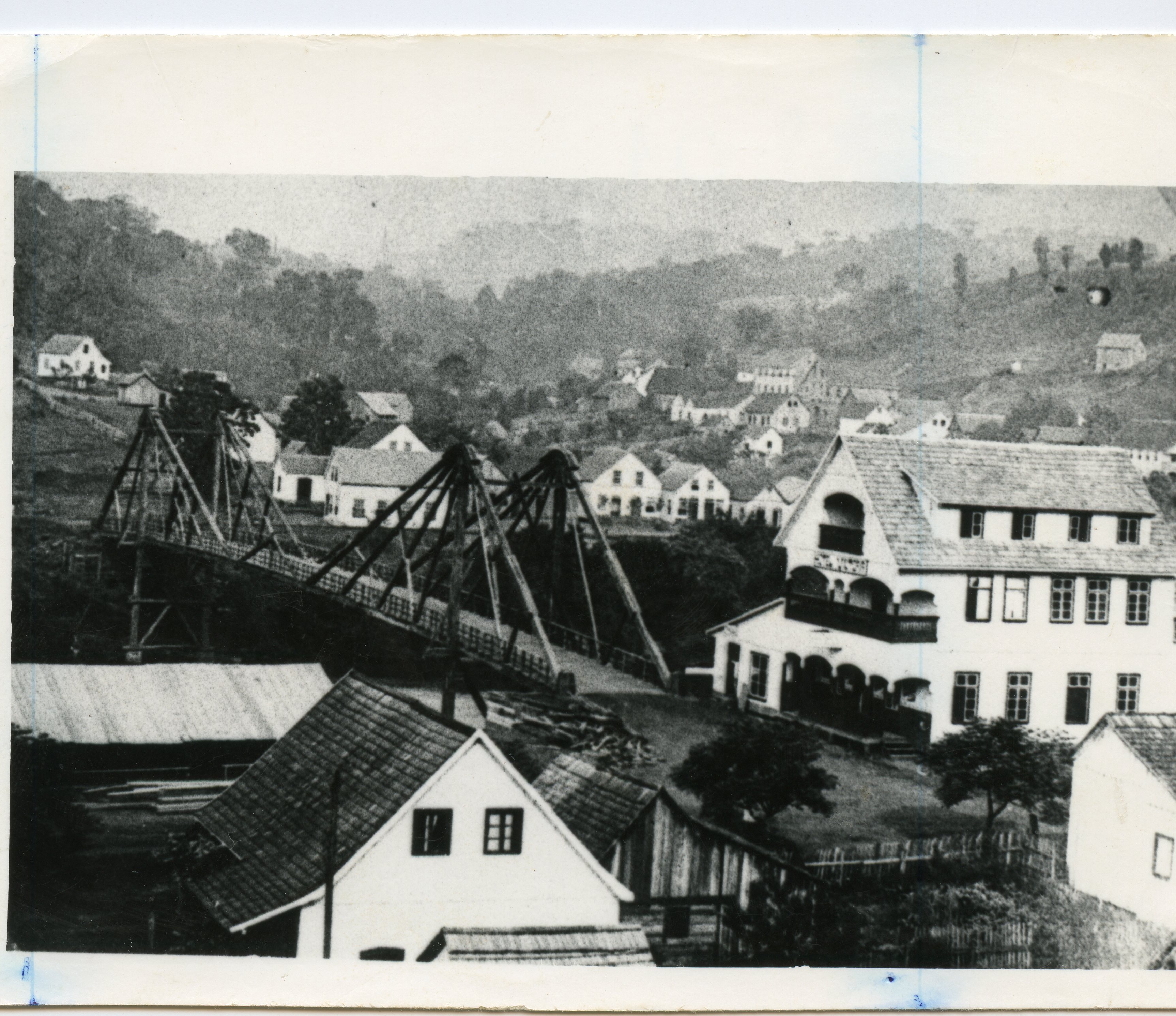 Fotografia em preto e branco de uma pequena cidade brasileira, possivelmente no início do século XX, destacando uma ponte pênsil de madeira sobre um rio e construções de estilo europeu. A paisagem rural ao fundo e a arquitetura sugerem um período de colonização, com casas de madeira e alvenaria típicas da época.