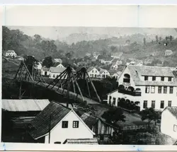 Fotografia em preto e branco de uma pequena cidade brasileira, possivelmente no início do século XX, destacando uma ponte pênsil de madeira sobre um rio e construções de estilo europeu. A paisagem rural ao fundo e a arquitetura sugerem um período de colonização, com casas de madeira e alvenaria típicas da época.