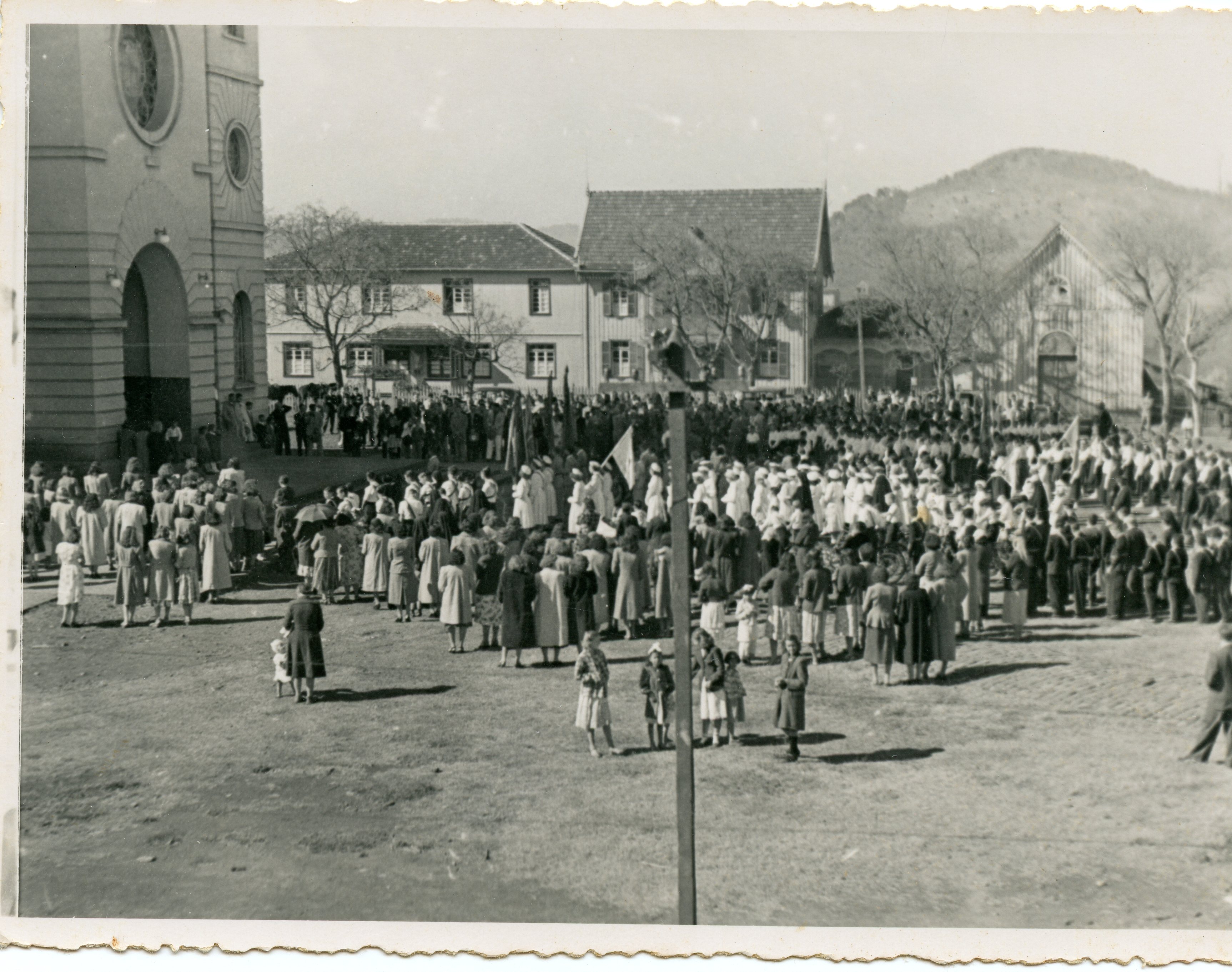 Uma fotografia em preto e branco que registra uma grande procissão ou evento religioso em frente a uma igreja de arquitetura clássica. A cena mostra uma multidão, incluindo muitas mulheres vestidas com roupas formais da época, sugerindo um contexto de colonização ou vida comunitária no interior do Brasil em meados do século XX.