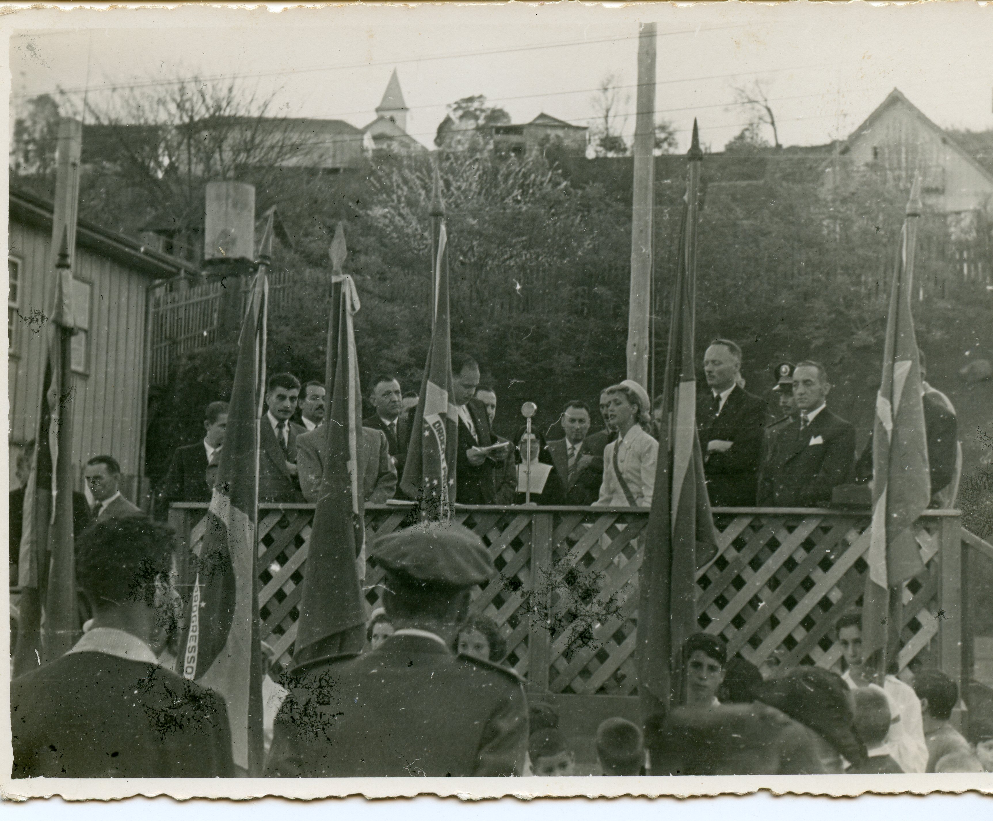 Fotografia em preto e branco de um evento cívico ou solene ao ar livre, possivelmente em uma pequena cidade brasileira, com autoridades discursando em um palanque de madeira. Observam-se bandeiras hasteadas, homens de terno e uma mulher ao microfone, com uma paisagem urbana simples e uma igreja ao fundo, sugerindo um contexto de meados do século XX.