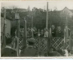 Fotografia em preto e branco de um evento cívico ou solene ao ar livre, possivelmente em uma pequena cidade brasileira, com autoridades discursando em um palanque de madeira. Observam-se bandeiras hasteadas, homens de terno e uma mulher ao microfone, com uma paisagem urbana simples e uma igreja ao fundo, sugerindo um contexto de meados do século XX.