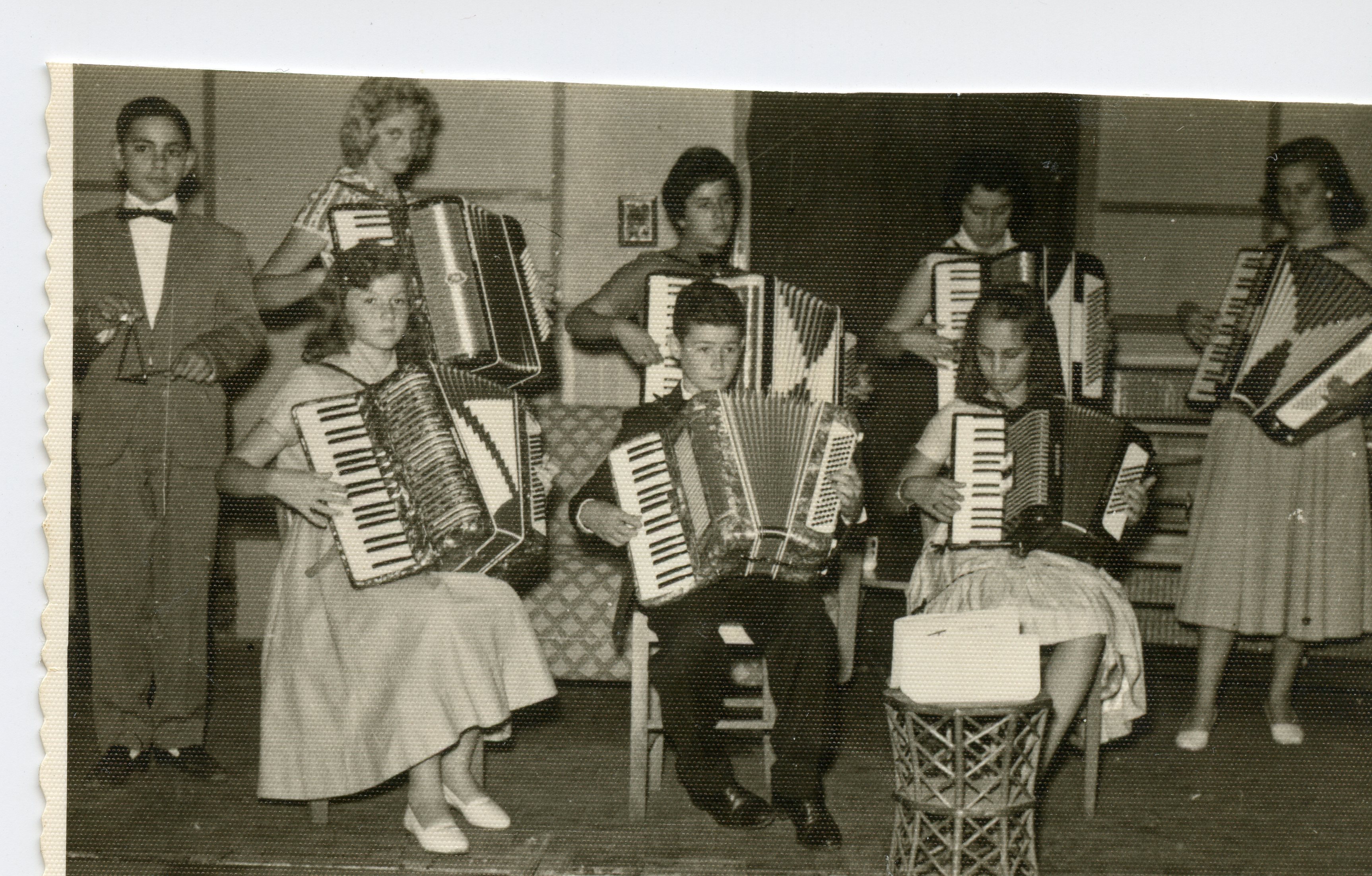 Uma fotografia em preto e branco retratando um grupo de jovens músicos tocando acordeons em um ambiente interno, possivelmente um recital ou apresentação escolar. O estilo das vestimentas, com vestidos rodados e ternos formais, sugere um contexto social de meados do século XX, comum em pequenas cidades brasileiras.