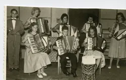 Uma fotografia em preto e branco retratando um grupo de jovens músicos tocando acordeons em um ambiente interno, possivelmente um recital ou apresentação escolar. O estilo das vestimentas, com vestidos rodados e ternos formais, sugere um contexto social de meados do século XX, comum em pequenas cidades brasileiras.