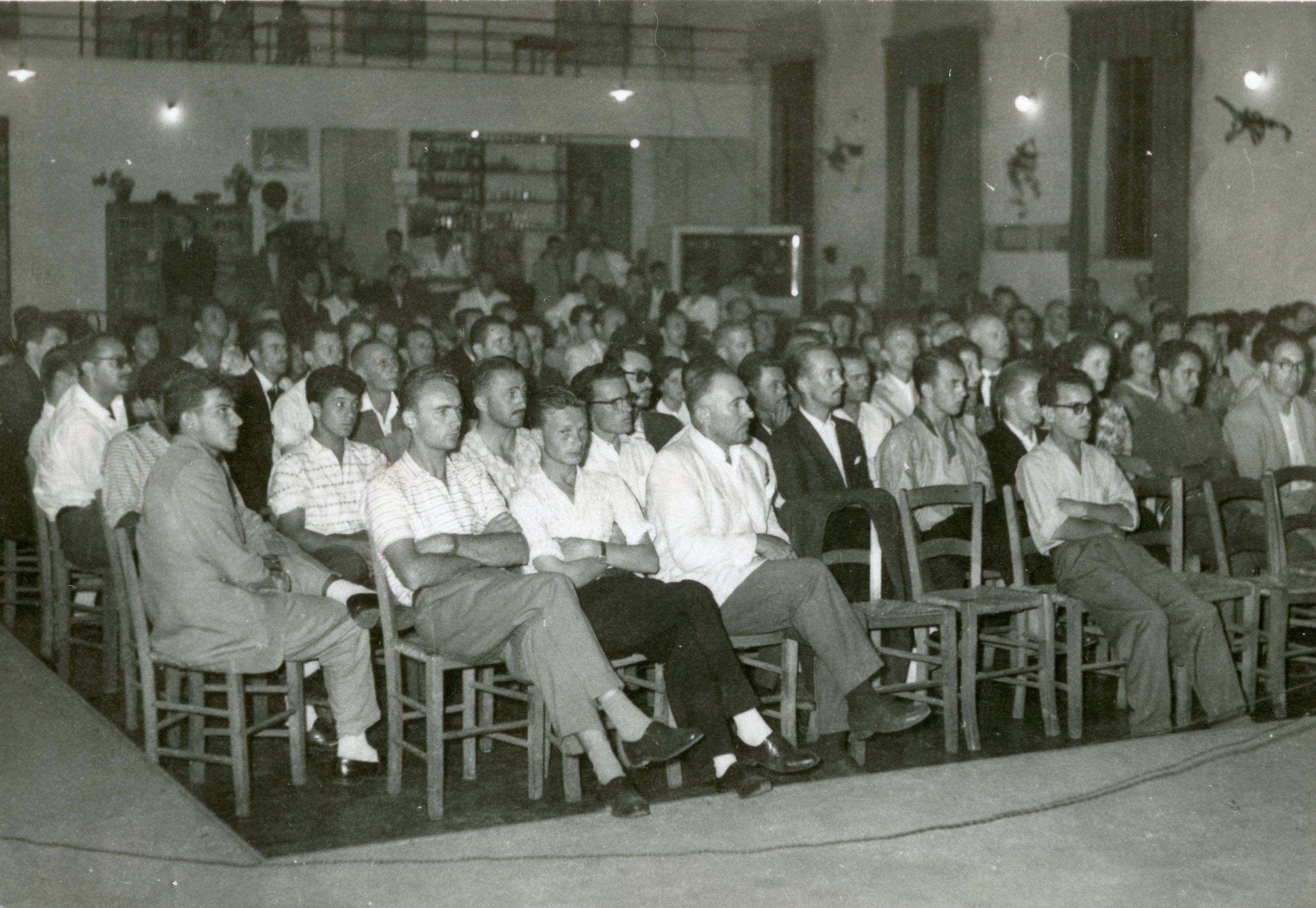 Uma fotografia em preto e branco que retrata uma plateia masculina numerosa sentada em cadeiras de madeira em um salão amplo. O ambiente sugere uma reunião comunitária, palestra ou evento social, típico de meados do século XX em uma cidade brasileira.