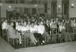 Uma fotografia em preto e branco que retrata uma plateia masculina numerosa sentada em cadeiras de madeira em um salão amplo. O ambiente sugere uma reunião comunitária, palestra ou evento social, típico de meados do século XX em uma cidade brasileira.