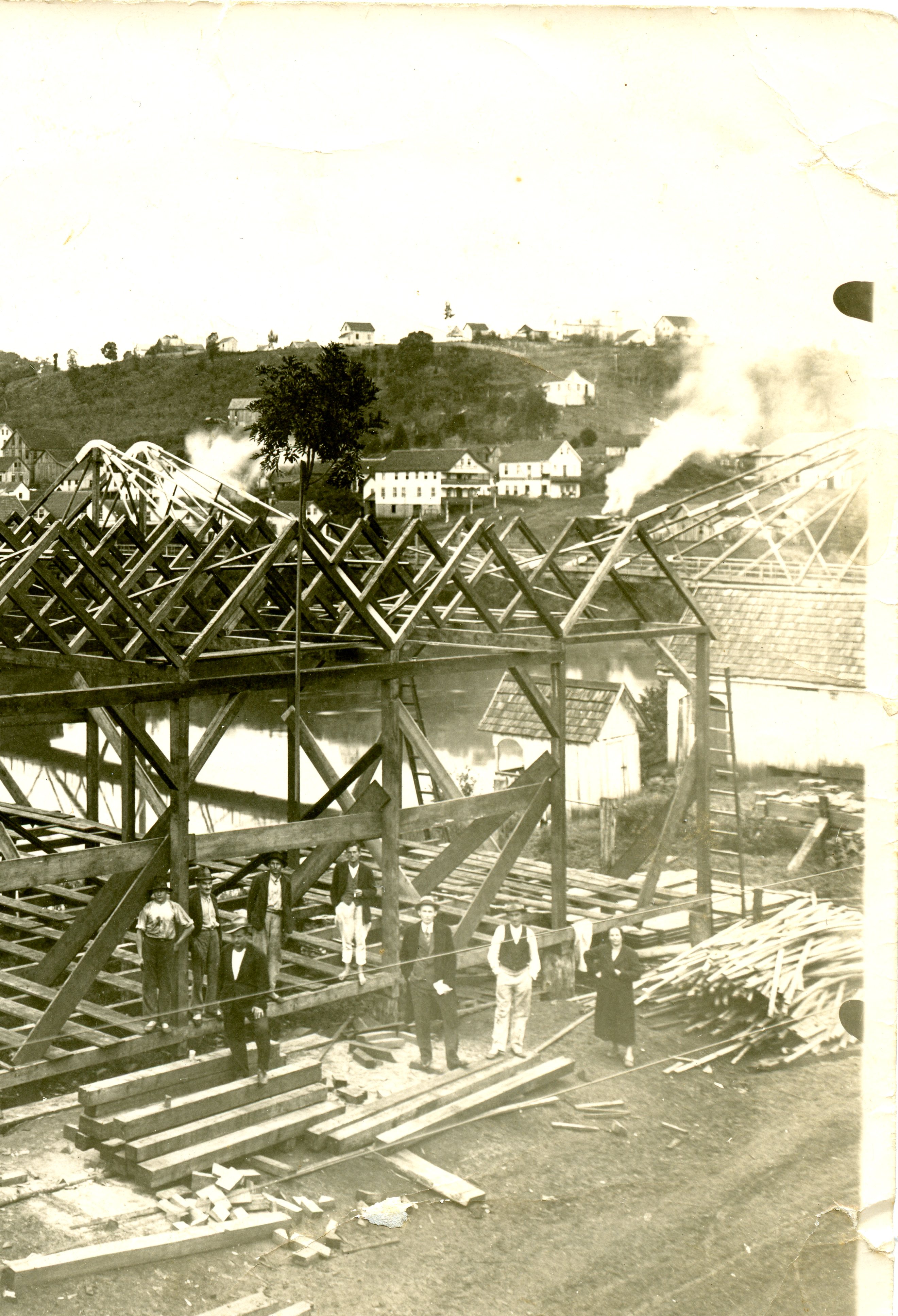 Fotografia histórica em preto e branco mostrando a construção de uma estrutura de madeira, possivelmente um galpão ou serraria, com um grupo de homens e uma mulher posando à frente. Ao fundo, observa-se uma paisagem rural com casas em uma colina, sugerindo o início do século XX em uma região de colonização no Brasil.