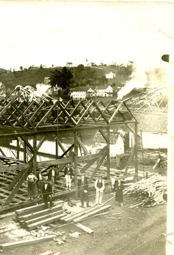 Fotografia histórica em preto e branco mostrando a construção de uma estrutura de madeira, possivelmente um galpão ou serraria, com um grupo de homens e uma mulher posando à frente. Ao fundo, observa-se uma paisagem rural com casas em uma colina, sugerindo o início do século XX em uma região de colonização no Brasil.