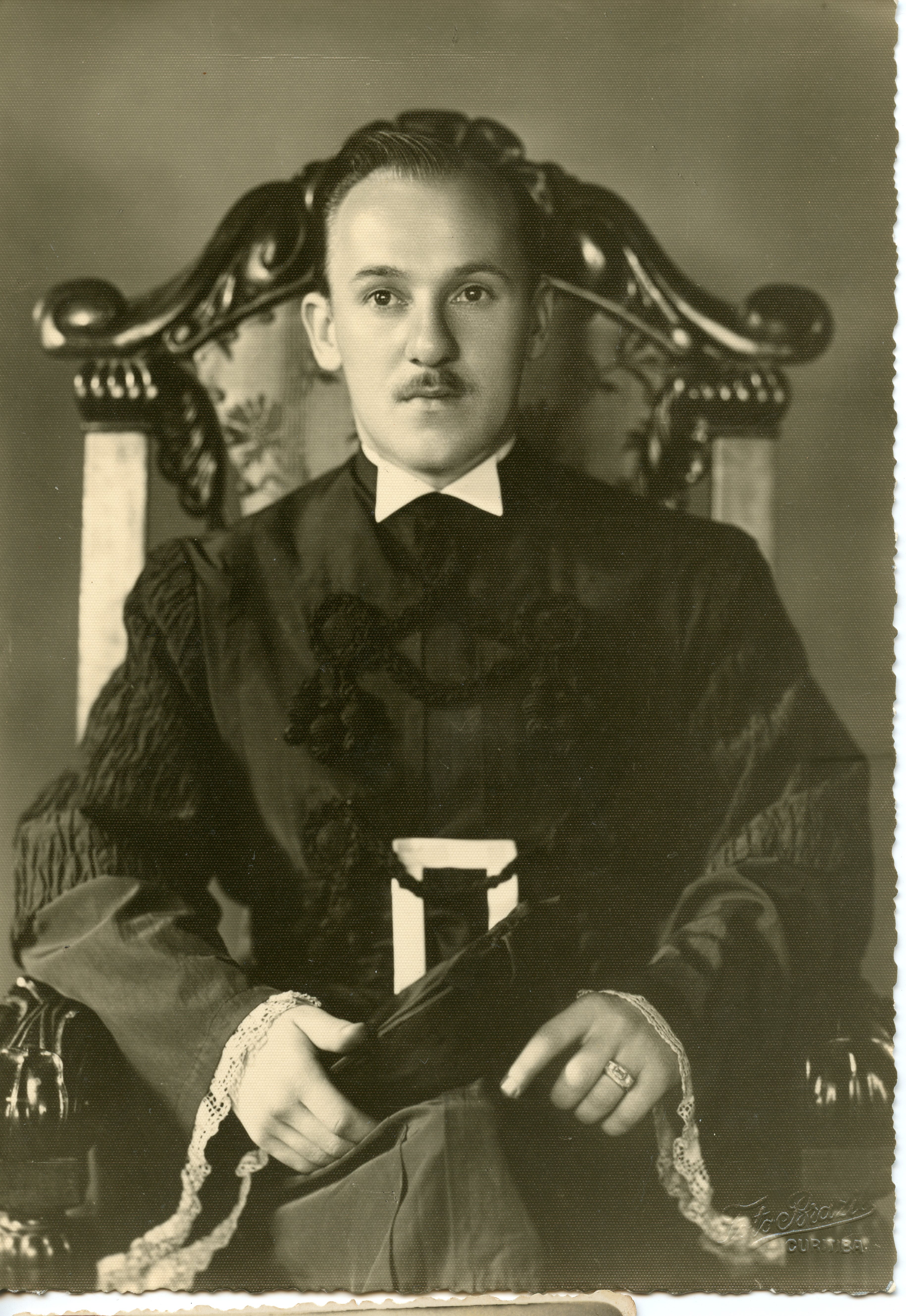 Retrato em estúdio de um homem jovem vestindo uma beca acadêmica formal, com detalhes em renda nos punhos e gola branca. A imagem, provavelmente de meados do século XX, sugere uma formatura ou evento acadêmico solene, com o indivíduo sentado em uma cadeira ornamentada.