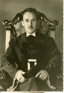 Retrato em estúdio de um homem jovem vestindo uma beca acadêmica formal, com detalhes em renda nos punhos e gola branca. A imagem, provavelmente de meados do século XX, sugere uma formatura ou evento acadêmico solene, com o indivíduo sentado em uma cadeira ornamentada.
