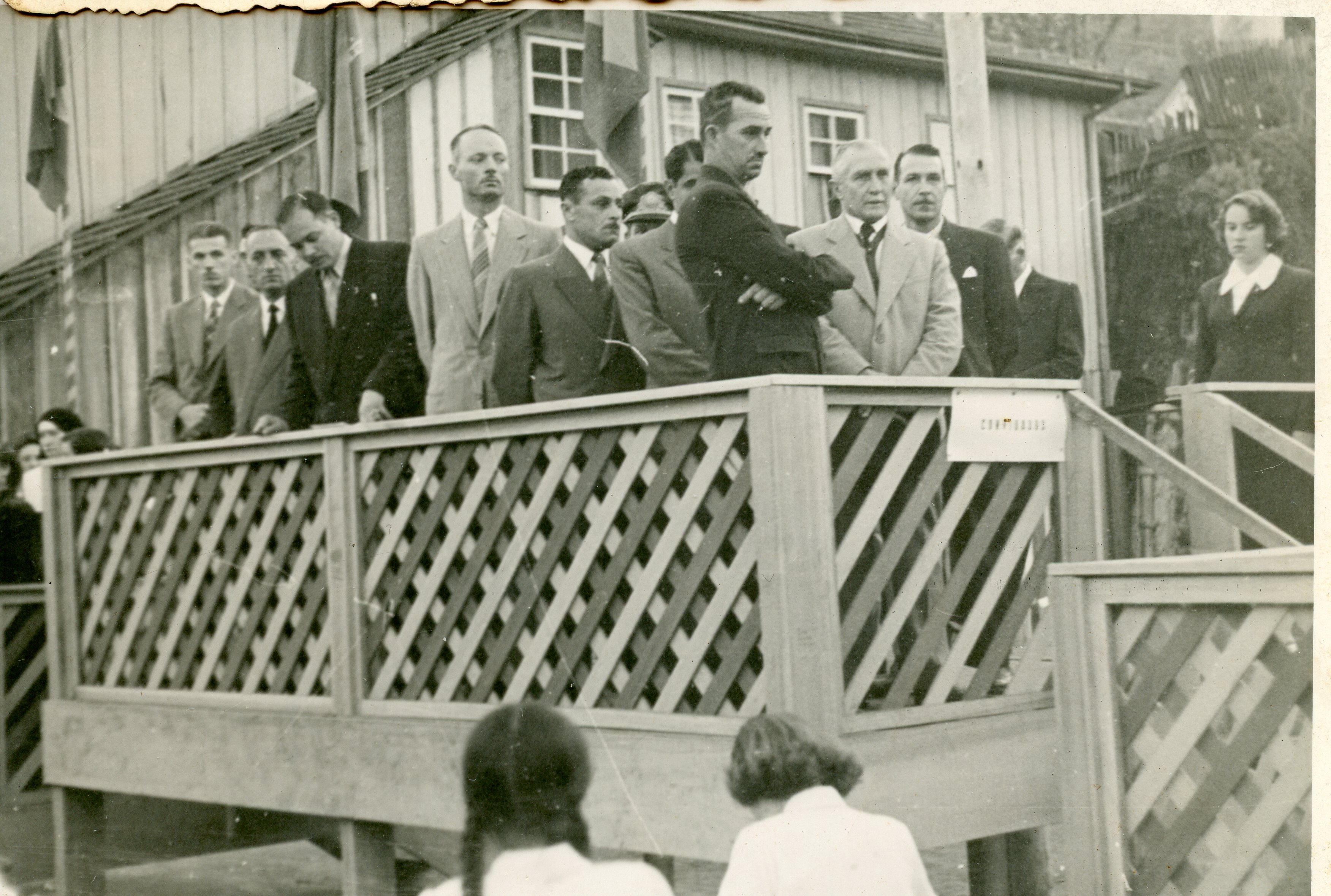 Uma fotografia em preto e branco que retrata um grupo de homens e uma mulher em um palanque de madeira, possivelmente durante uma cerimônia cívica ou evento público. O estilo das vestimentas, com ternos e gravatas, sugere um contexto formal típico da metade do século XX em uma cidade brasileira em desenvolvimento.