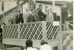 Uma fotografia em preto e branco que retrata um grupo de homens e uma mulher em um palanque de madeira, possivelmente durante uma cerimônia cívica ou evento público. O estilo das vestimentas, com ternos e gravatas, sugere um contexto formal típico da metade do século XX em uma cidade brasileira em desenvolvimento.