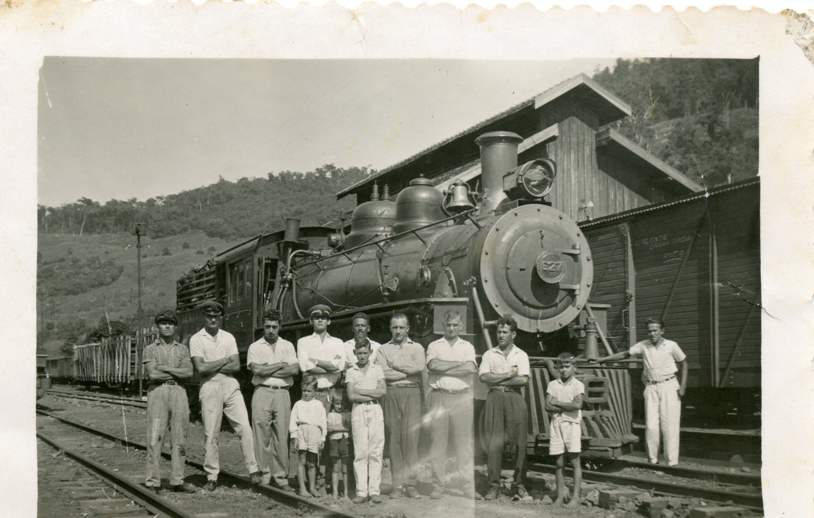 Fotografia em preto e branco retratando um grupo de homens e crianças posando em frente a uma locomotiva a vapor em uma estação ferroviária. A cena sugere um contexto de trabalho ou cotidiano ferroviário no interior do Brasil, possivelmente na primeira metade do século XX, com vestimentas típicas da época.