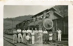 Fotografia em preto e branco retratando um grupo de homens e crianças posando em frente a uma locomotiva a vapor em uma estação ferroviária. A cena sugere um contexto de trabalho ou cotidiano ferroviário no interior do Brasil, possivelmente na primeira metade do século XX, com vestimentas típicas da época.