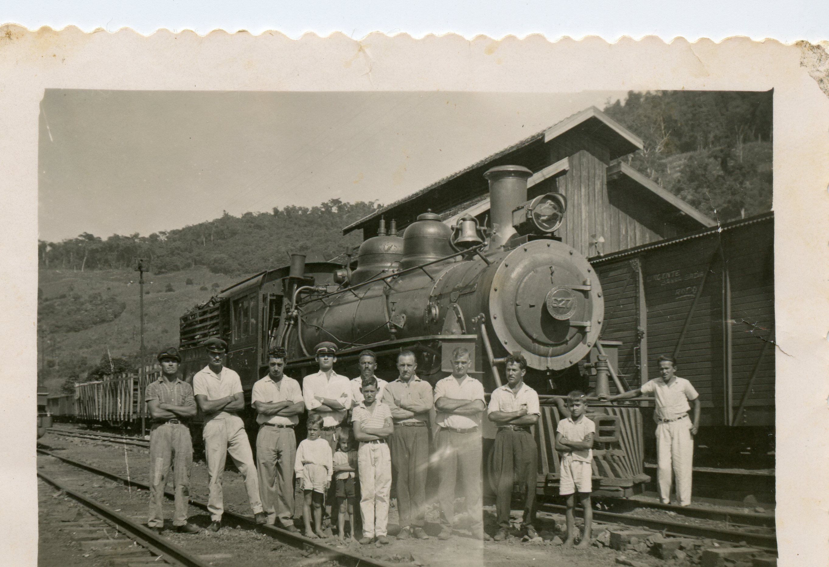 Fotografia em preto e branco retratando um grupo de homens e crianças posando em frente a uma locomotiva a vapor em uma estação ferroviária. A cena sugere o contexto de expansão ferroviária no Brasil, possivelmente em meados do século XX, com vestimentas típicas da época e uma estrutura de madeira ao fundo.