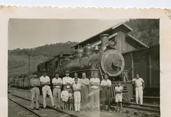 Fotografia em preto e branco retratando um grupo de homens e crianças posando em frente a uma locomotiva a vapor em uma estação ferroviária. A cena sugere o contexto de expansão ferroviária no Brasil, possivelmente em meados do século XX, com vestimentas típicas da época e uma estrutura de madeira ao fundo.
