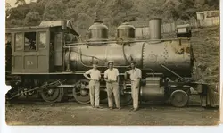 Fotografia em preto e branco mostrando três trabalhadores ferroviários posando ao lado de uma locomotiva a vapor numerada '10' e identificada como 'PARANA'. A cena sugere o início do século XX, refletindo a expansão das ferrovias no Brasil, com os homens vestindo roupas de trabalho típicas da época em um ambiente rural ou de estação ferroviária.