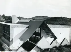 Fotografia em preto e branco capturando uma estrutura arquitetônica moderna com telhados angulares e geométricos, possivelmente em fase de construção ou finalização. O cenário ao fundo revela uma paisagem rural ou de transição urbana, sugerindo um projeto arquitetônico arrojado em um contexto regional brasileiro.
