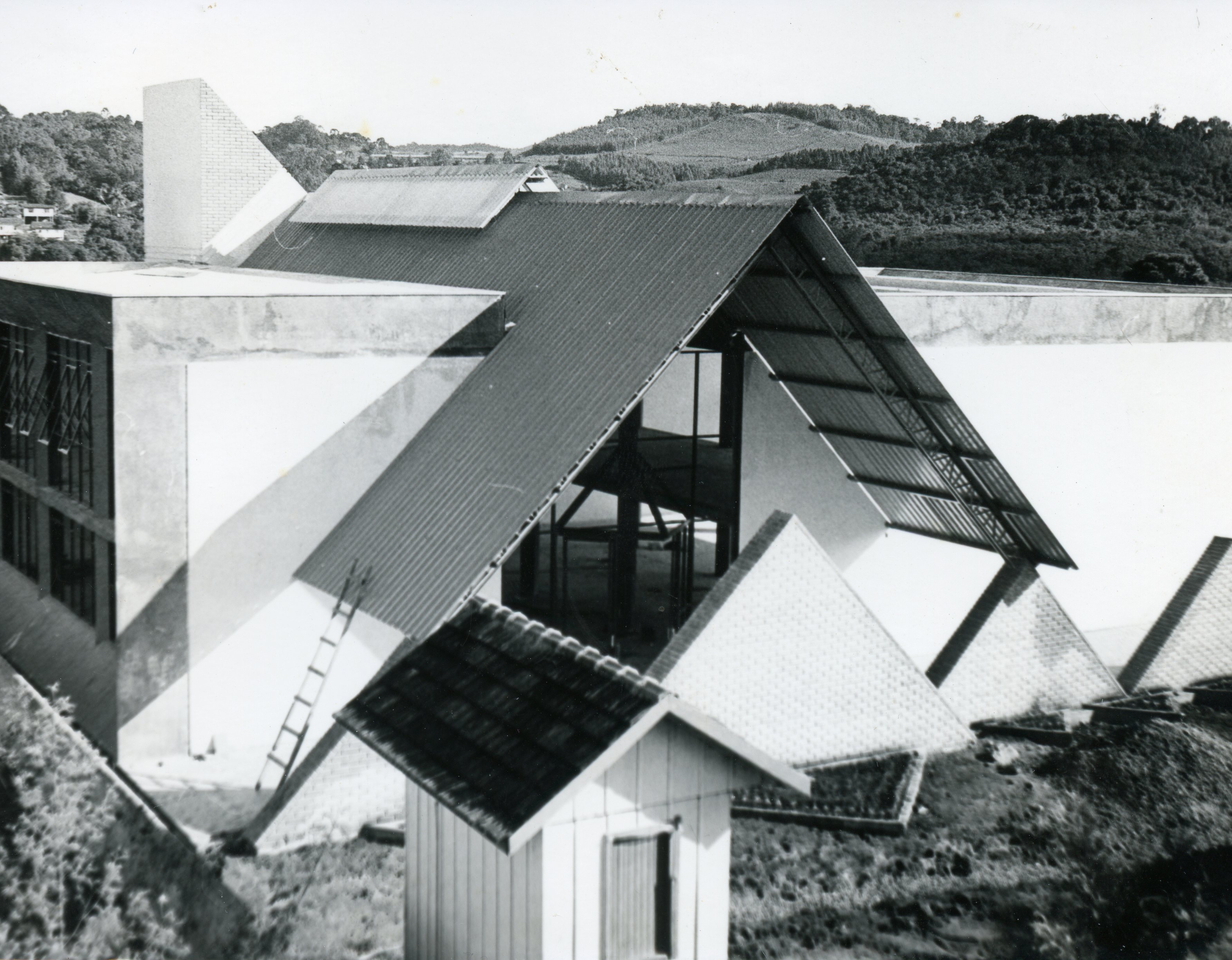 Fotografia em preto e branco mostrando a construção de uma edificação com arquitetura moderna, caracterizada por telhados em formato triangular e linhas geométricas acentuadas. O cenário sugere uma área em desenvolvimento, possivelmente em uma região serrana ou rural, com uma pequena estrutura de madeira em primeiro plano e colinas ao fundo.
