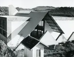 Fotografia em preto e branco mostrando a construção de uma edificação com arquitetura moderna, caracterizada por telhados em formato triangular e linhas geométricas acentuadas. O cenário sugere uma área em desenvolvimento, possivelmente em uma região serrana ou rural, com uma pequena estrutura de madeira em primeiro plano e colinas ao fundo.