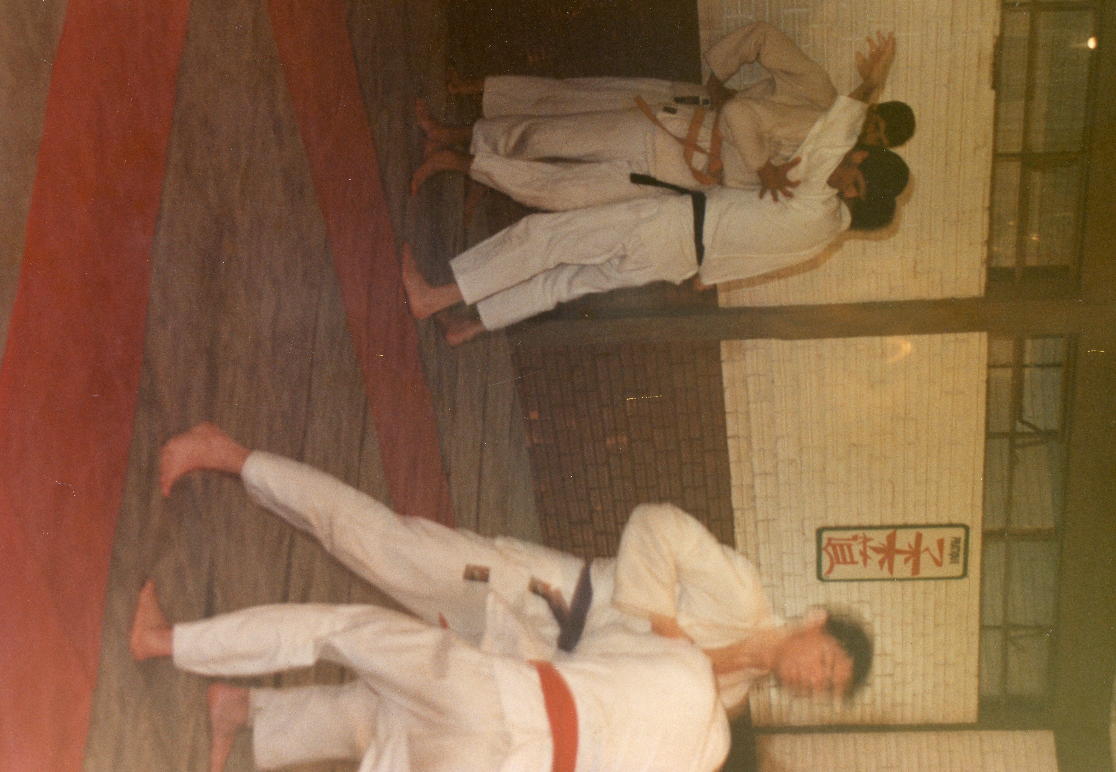 Uma fotografia colorida que captura uma sessão de treinamento de artes marciais, possivelmente judô, em um ambiente interno. Os praticantes vestem quimonos brancos com faixas de diferentes cores, realizando técnicas de projeção sobre um tatame, com uma placa com caracteres orientais visível na parede ao fundo.
