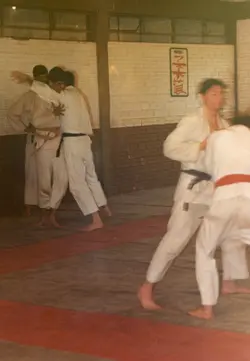 Uma fotografia colorida que captura uma sessão de treinamento de artes marciais, possivelmente judô, em um ambiente interno. Os praticantes vestem quimonos brancos com faixas de diferentes cores, realizando técnicas de projeção sobre um tatame, com uma placa com caracteres orientais visível na parede ao fundo.