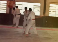 Uma fotografia de época capturando um treino de artes marciais, possivelmente judô, em um ambiente interno com janelas amplas. Os praticantes vestem quimonos brancos e faixas de diferentes cores, sugerindo uma prática esportiva em um ginásio ou academia local.