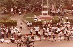 Uma cena ao ar livre em uma praça pública, provavelmente durante um evento escolar ou feira cultural, com diversas crianças e adolescentes vestindo uniformes escolares. A imagem apresenta elementos típicos das décadas de 1970 ou 1980, como o estilo das roupas, a motocicleta e a estética da fotografia, sugerindo um ambiente comunitário em uma cidade brasileira.