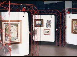 Uma exposição de arte organizada em painéis brancos suspensos por uma estrutura metálica treliçada de cor vermelha. A cena apresenta diversas pinturas emolduradas, sugerindo um evento cultural ou galeria de arte contemporânea.