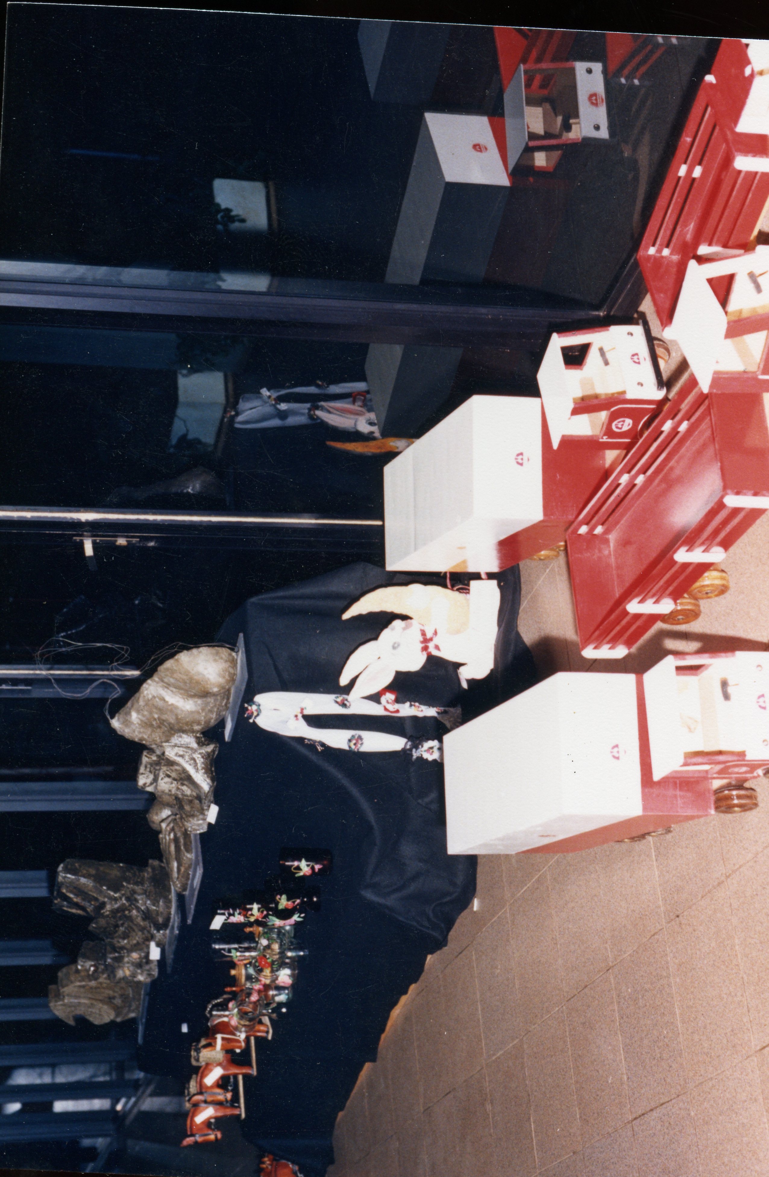 Uma fotografia que mostra uma vitrine ou exposição interna contendo brinquedos de madeira artesanais, incluindo trens e figuras decorativas como um coelho. A cena parece ser de uma exposição comercial ou escolar, possivelmente datada entre as décadas de 1980 e 1990, com elementos decorativos dispostos sobre um tecido escuro.