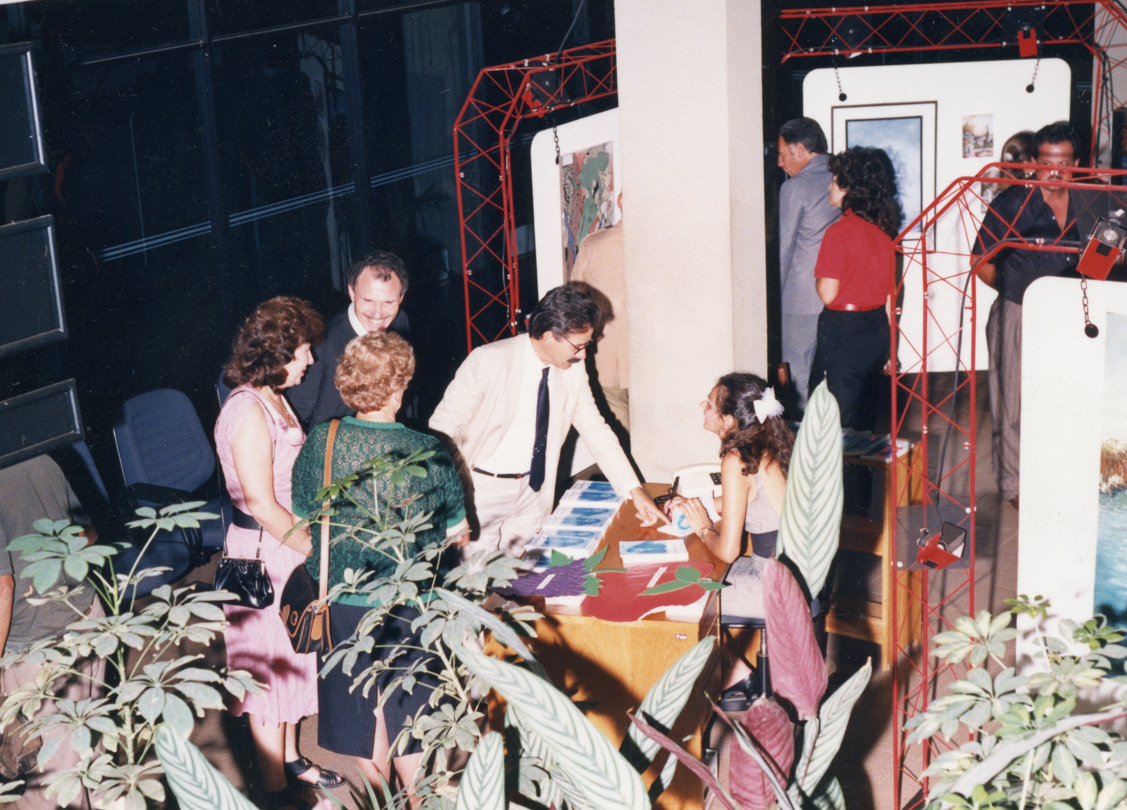 Uma cena de evento social ou cultural, possivelmente uma exposição de arte ou lançamento de livros, realizada em um ambiente interno com estruturas metálicas vermelhas. As pessoas estão vestidas com moda típica do final da década de 1980 ou início da de 1990, interagindo em torno de uma mesa de recepção.