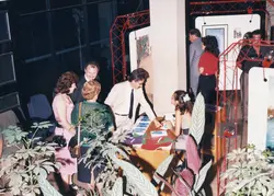 Uma cena de evento social ou cultural, possivelmente uma exposição de arte ou lançamento de livros, realizada em um ambiente interno com estruturas metálicas vermelhas. As pessoas estão vestidas com moda típica do final da década de 1980 ou início da de 1990, interagindo em torno de uma mesa de recepção.