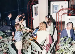Uma cena de vernissage ou exposição de arte em um ambiente interno, com pessoas vestidas de forma social conversando e observando quadros dispostos em estruturas metálicas vermelhas. A estética das roupas e a qualidade da fotografia sugerem um evento cultural ocorrido provavelmente na década de 1980 ou início de 1990.