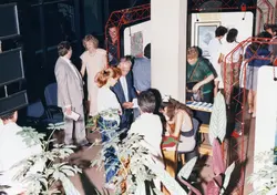Uma fotografia de um evento social ou exposição de arte em um ambiente interno, possivelmente na década de 1980 ou início de 1990. Pessoas vestidas com trajes formais e casuais da época interagem em um espaço decorado com estruturas metálicas vermelhas e quadros, sugerindo uma vernissage ou recepção cultural.