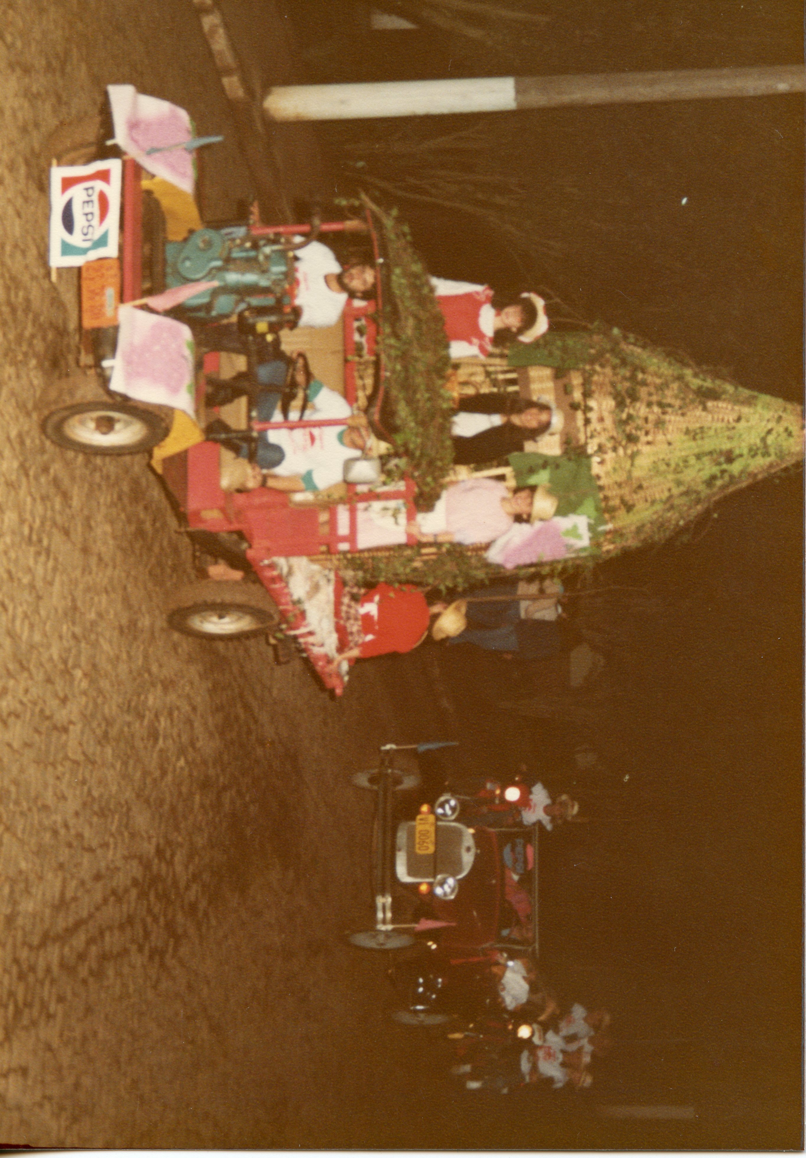 Fotografia noturna de um desfile ou evento festivo, possivelmente em uma cidade do interior, apresentando um veículo decorado com folhagens e pessoas a bordo. O estilo das roupas e a qualidade da imagem sugerem um registro das décadas de 1980 ou 1990, com a presença de um logotipo da Pepsi e um veículo antigo seguindo o carro principal.