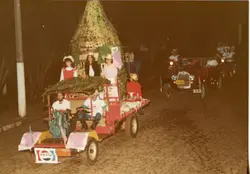 Fotografia noturna de um desfile ou evento festivo, possivelmente em uma cidade do interior, apresentando um veículo decorado com folhagens e pessoas a bordo. O estilo das roupas e a qualidade da imagem sugerem um registro das décadas de 1980 ou 1990, com a presença de um logotipo da Pepsi e um veículo antigo seguindo o carro principal.