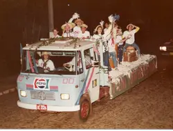 Uma fotografia noturna de um caminhão decorado como carro alegórico, transportando um grupo de jovens com chapéus de palha e roupas informais. O veículo, um Mercedes-Benz, exibe a frase 'Veni Tutti Bever Vino' na lateral, sugerindo uma celebração cultural ou festividade típica de colônias italianas no sul do Brasil.