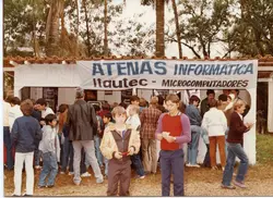 Uma fotografia colorida que registra um estande da 'Atenas Informática' promovendo microcomputadores Itautec, provavelmente em uma feira ou evento público ao ar livre. A cena mostra diversas pessoas, incluindo muitas crianças e jovens, vestindo roupas típicas da década de 1980, em um ambiente que sugere uma pequena cidade brasileira durante a popularização da computação pessoal.