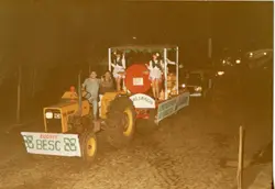 Uma fotografia noturna de um desfile ou evento festivo, possivelmente em uma cidade do interior, apresentando um trator Valmet decorado como carro alegórico. O veículo carrega pessoas vestidas com trajes típicos, sugerindo uma celebração cultural ou agrícola, com faixas da equipe BESC e referências aos 'Italianos'.