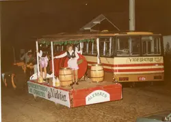 Uma fotografia noturna de um desfile temático em Videira, Santa Catarina, exibindo um carro alegórico puxado por um trator. O veículo, da empresa Videiratur, está decorado com barris de madeira e faixas que celebram as origens alemãs da colonização local, com pessoas vestidas com trajes típicos.