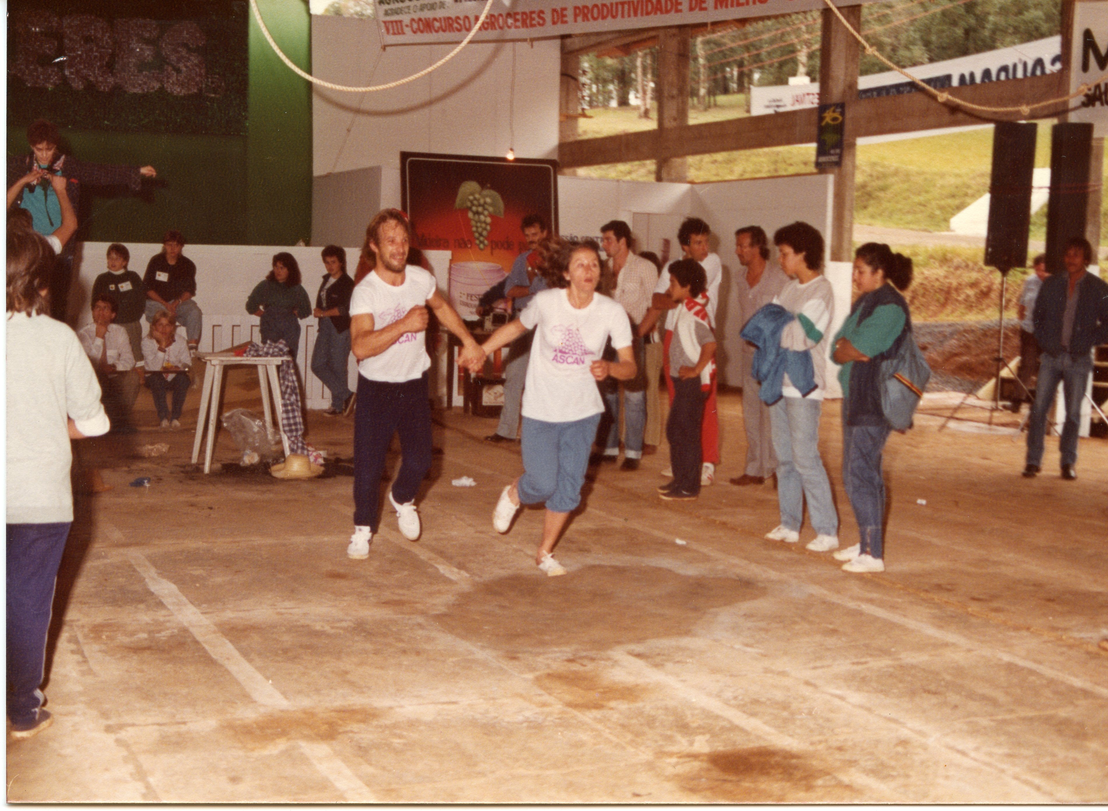 Uma cena de um evento comunitário, possivelmente uma gincana ou competição esportiva, realizada em um pavilhão coberto. O ambiente sugere um contexto rural ou de interior, com cartazes mencionando concursos agrícolas e festividades, típicos de eventos regionais brasileiros das décadas de 1980 ou 1990.