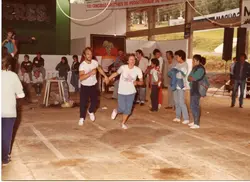 Uma cena de um evento comunitário, possivelmente uma gincana ou competição esportiva, realizada em um pavilhão coberto. O ambiente sugere um contexto rural ou de interior, com cartazes mencionando concursos agrícolas e festividades, típicos de eventos regionais brasileiros das décadas de 1980 ou 1990.