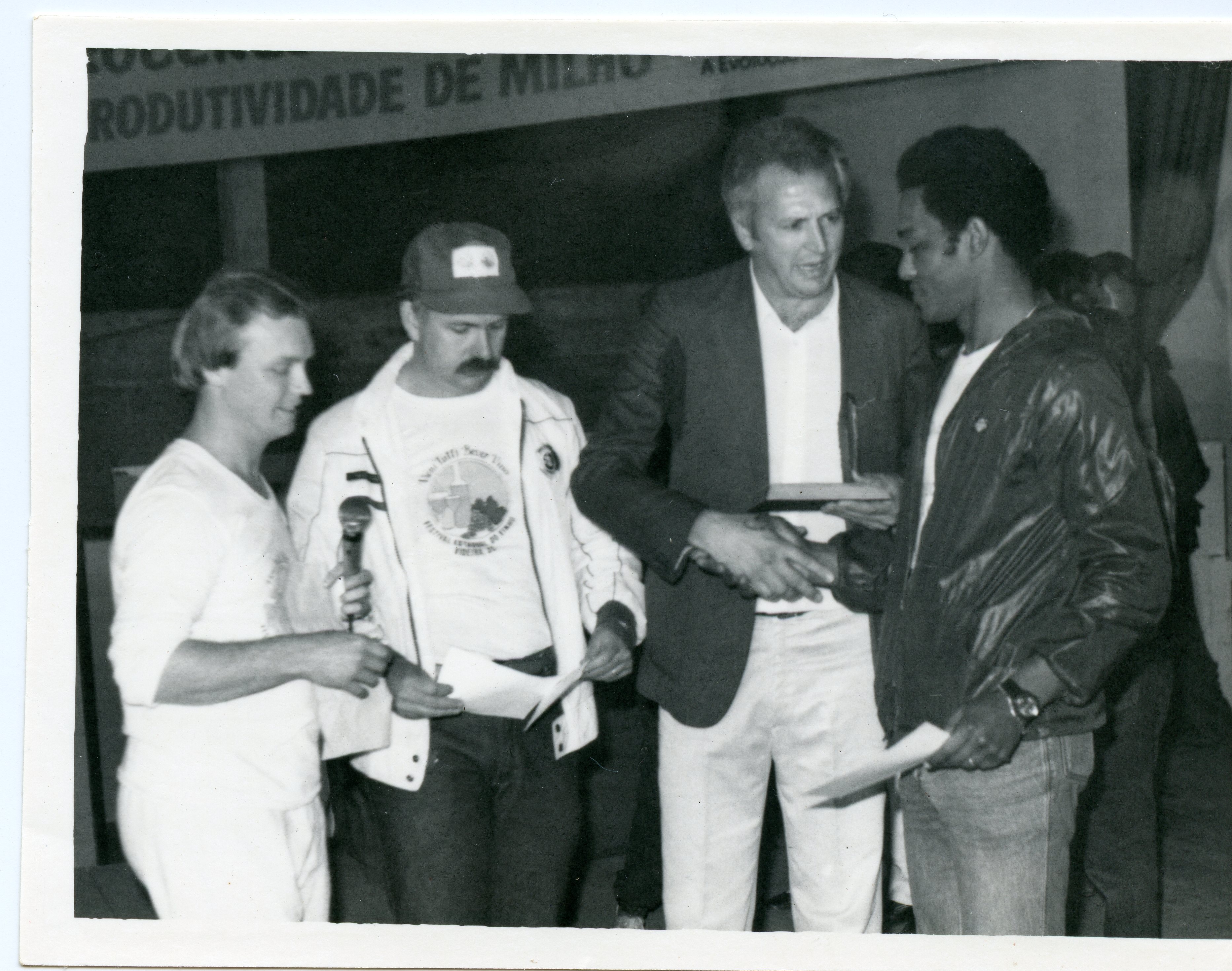 Fotografia em preto e branco registrando uma cerimônia de premiação ou evento comunitário, possivelmente relacionado à agricultura, dado o cartaz ao fundo mencionando 'produtividade de milho'. Três homens aparecem em destaque, com um deles cumprimentando o outro, sugerindo uma entrega de certificados ou troféus em um ambiente interno, típico de eventos sociais ou rurais da década de 1980.