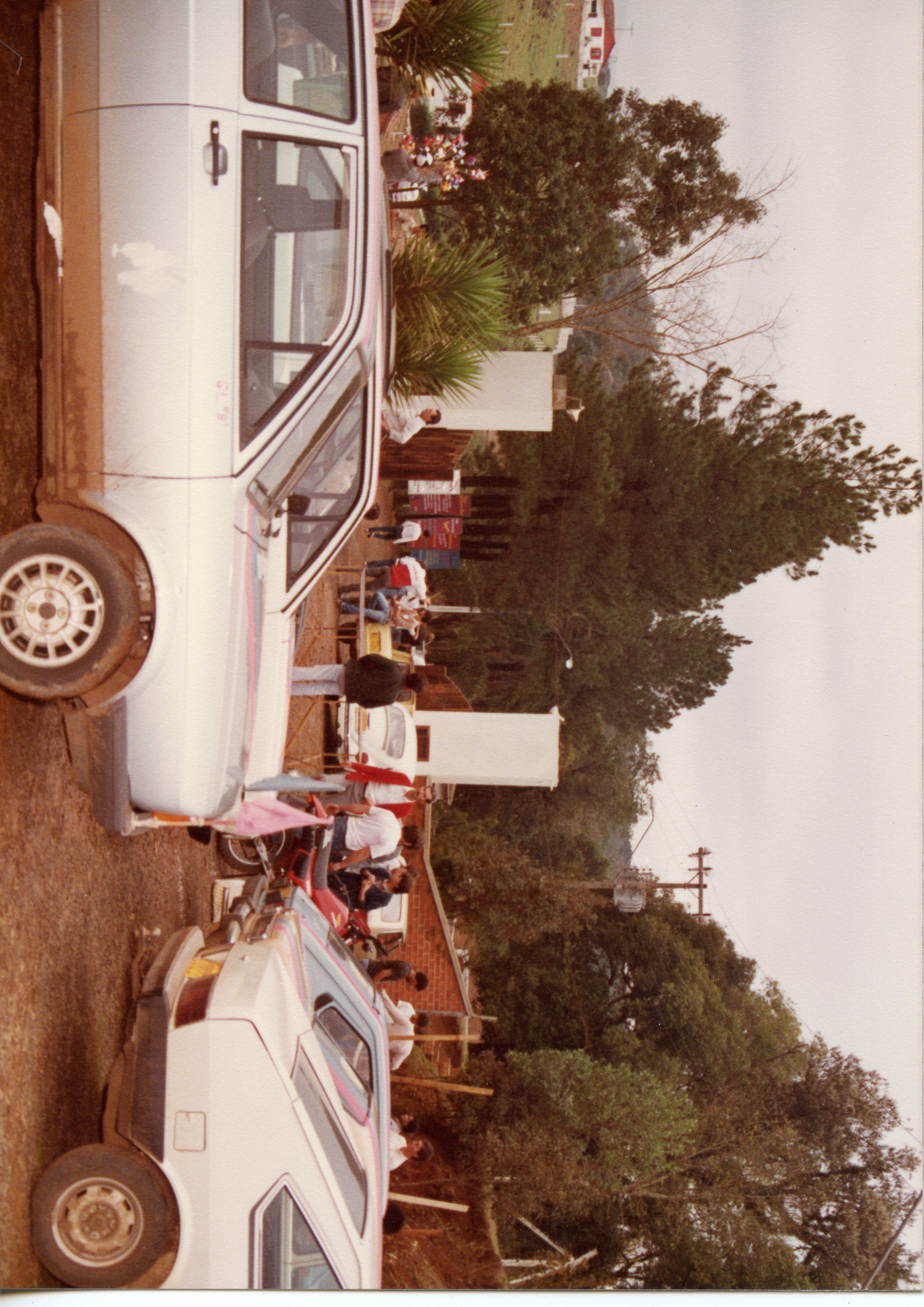 Uma fotografia colorida de época, provavelmente das décadas de 1980 ou 1990, mostrando um grupo de pessoas reunidas em uma área externa próxima a veículos estacionados. O cenário sugere um ambiente rural ou semi-urbano com vegetação e construções simples, comum em pequenas cidades brasileiras.